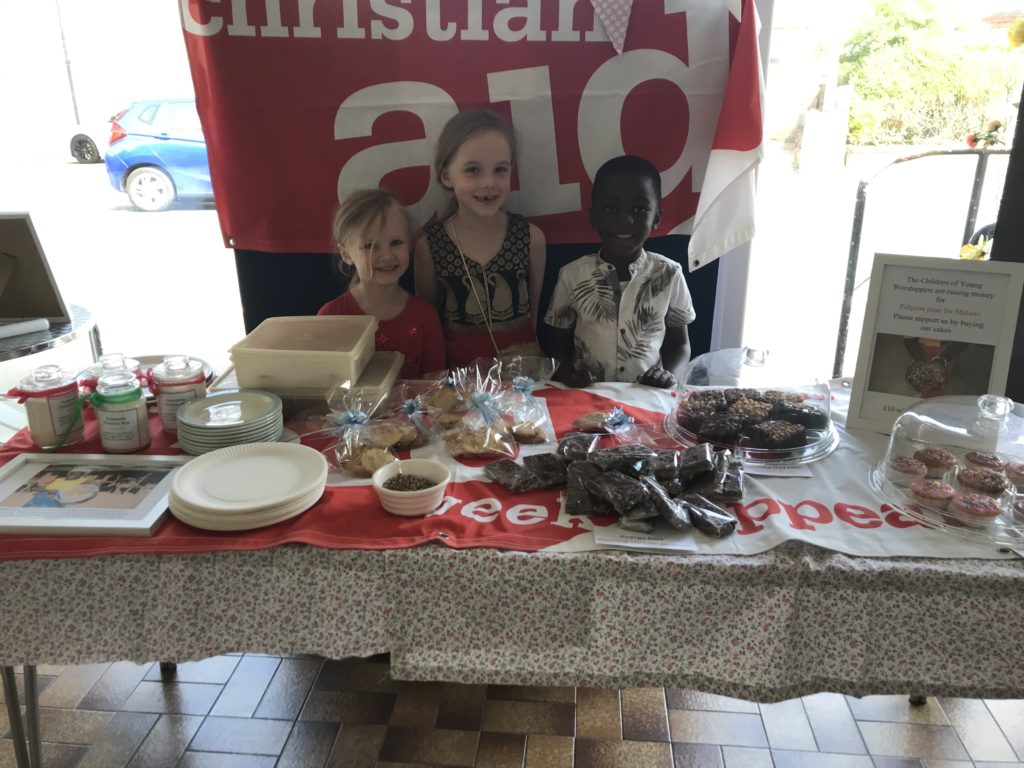 image of three smiling children behind a table full of cakes. a christian aid banner hangs behind the children